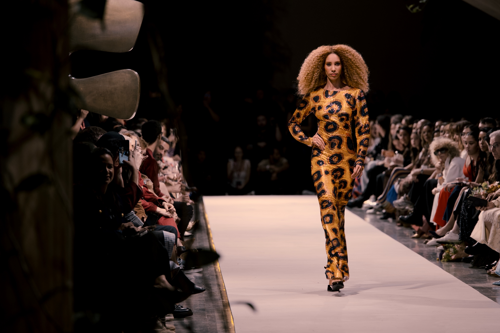 Fashion runway show with a model walking the catwalk in a leopard-print outfit while an audience watches from both sides